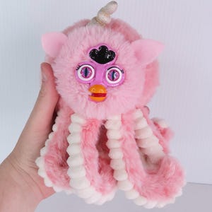 Könnte beinhalten: Ein rosa Plüschtier mit einem Furby-ähnlichen Gesicht und Tentakeln im Oktopus-Stil. Das Spielzeug hat große, runde Augen mit lila und blauen Details, eine schwarze Krone und ein goldenes Horn. Die Tentakel sind rosa und weiß.