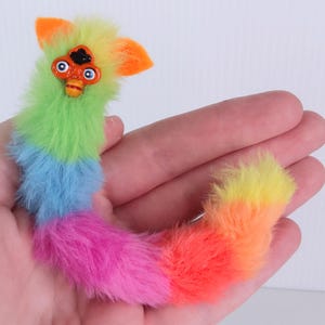 May include: A colorful plush toy in the shape of a creature, held in a hand. The toy features a rainbow gradient of colors, including green, blue, pink, orange, and yellow. It has a detailed face with orange and black accents.