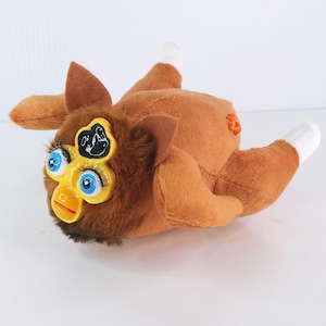 May include: A brown plush toy with large blue eyes, long eyelashes, and a yellow beak. The toy has brown ears and limbs, with white tips on the feet. A black and white design is on the forehead. The toy is lying on its back.