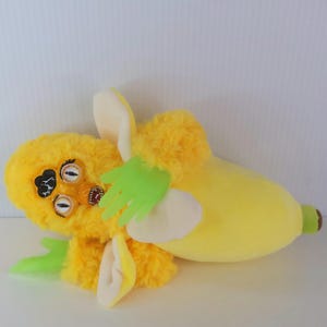 May include: A banana-shaped plush toy with a yellow body and a furry yellow head featuring three eyes and a mouth. The toy has green hands and white accents.