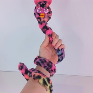 May include: A plush toy in the shape of a Furby with a pink, black, and orange leopard print design. The Furby has pink ears, purple eyes, and an orange beak. The toy is held in a hand, with the tail wrapped around the wrist.