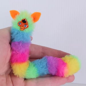May include: A colorful, plush toy in the shape of a caterpillar. The toy features a rainbow of colors, including yellow, pink, purple, blue, and green. It has orange ears and a face with two blue eyes and a small orange mouth. The toy is soft and fluffy.