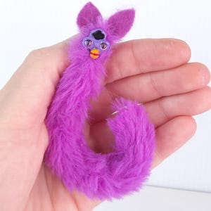 May include: A small, purple, plush toy in the shape of a Furby. The toy has a fuzzy body, large ears, and a face with eyes and a beak. The Furby is held in a person's hand.