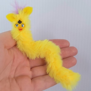May include: A yellow plush creature with a whimsical design. It has large blue eyes, a small orange beak, and a tuft of pink and purple feathers. The body is long and worm-like, covered in soft, yellow fur.