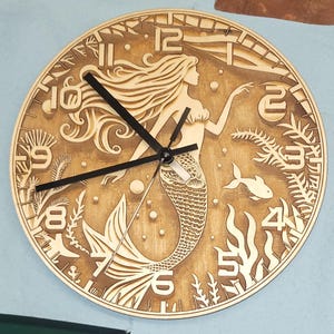 May include: A wooden clock with a mermaid design. The clock face features Roman numerals and a mermaid with flowing hair and a tail. The clock is surrounded by a decorative border of seaweed and bubbles.