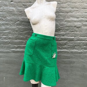 Pu&ograve; includere: Una minigonna verde con orlo a balze, due tasche e una chiusura a bottone decorativa. La gonna ha un abbellimento di gioielli bianchi sul lato destro.