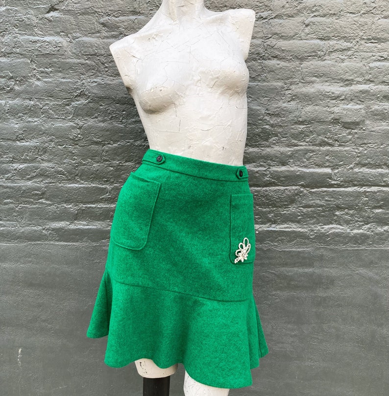 Pu&ograve; includere: Una minigonna verde con orlo a balze, due tasche e una chiusura a bottone decorativa. La gonna ha un abbellimento di gioielli bianchi sul lato destro.