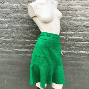 Pu&ograve; includere: Una gonna di lana verde con un orlo a balze e una sola tasca sul davanti. La gonna ha una chiusura a bottone in vita.
