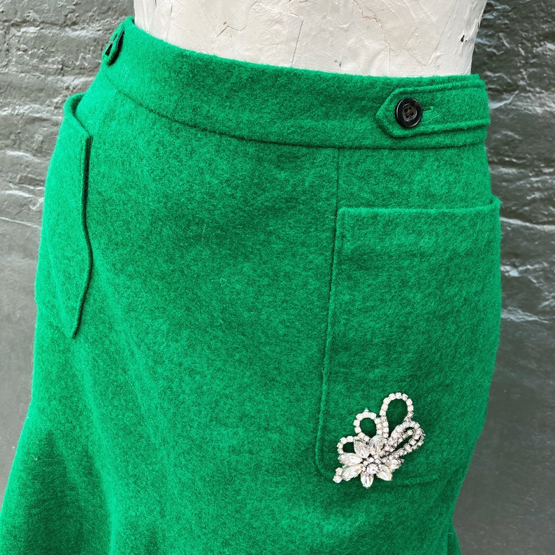 Pu&ograve; includere: Una gonna di lana verde con una grande tasca sul lato destro. La tasca &egrave; decorata con una spilla di strass.