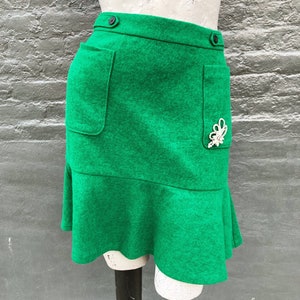 Pu&ograve; includere: Una minigonna verde con orlo a balze e due tasche. La gonna ha una chiusura a bottone e un abbellimento decorativo di perle.