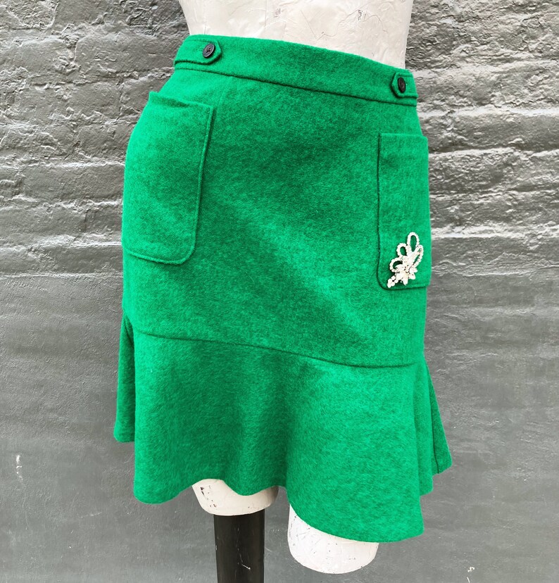 Pu&ograve; includere: Una minigonna verde con orlo a balze e due tasche. La gonna ha una chiusura a bottone e un abbellimento decorativo di perle.