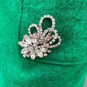 Pu&ograve; includere: Una spilla di strass argentata con un motivo floreale su uno sfondo di tessuto verde.