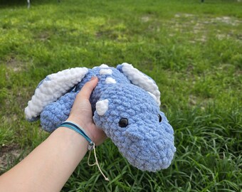 May include: A light blue crocheted dinosaur with white accents and black eyes. The handmade plush toy features a textured surface and is held against a backdrop of green grass.