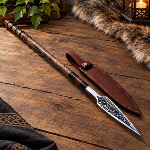 May include: A long spear featuring a dark wood shaft with black and red leather wrappings, paired with a sharp metal spearhead engraved with intricate patterns. A matching brown leather sheath with a brass snap button sits beside the spear.