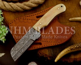 May include: A custom knife with a patterned blade and a cream-coloured handle. The knife is displayed on a brown leather sheath. The text "NHM BLADES Custom Hand Made Knives" is visible. The background includes a rope and decorative elements.
