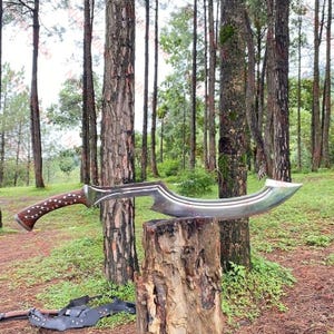 Puede incluir: Una hoja curva de color plateado con un mango de madera con un patrón marrón oscuro detallado. La hoja descansa sobre un tocón de árbol en un entorno forestal, mostrando su diseño único y artesanía.