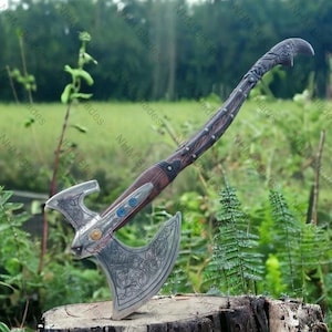 May include: A detailed Viking axe with a silver-colored head and a wooden handle. The axe head features intricate engraved designs and is adorned with blue and gold accents. The handle is dark brown with decorative metal studs. The axe is displayed outdoors.