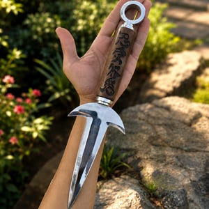 May include: A silver and brown kunai knife with a ring at the top. The handle is brown with Japanese characters etched into it. The blade is silver and has a unique shape. The knife is held in a hand against a blurred background of greenery and rocks.