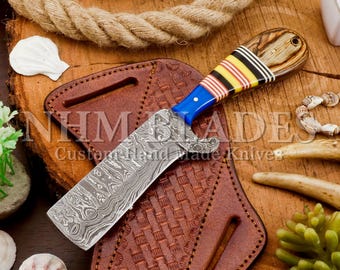 May include: A custom knife with a patterned blade and a handle with blue, yellow, and black stripes. It sits on a brown leather sheath with a woven design. The image includes the text "NHM Blades Custom Hand Made Knives".