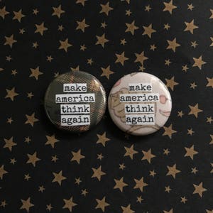 Può includere: Due bottoni rotondi con il testo "make america think again" stampato su di essi. Un bottone ha un motivo a quadri e l'altro ha un motivo floreale.
