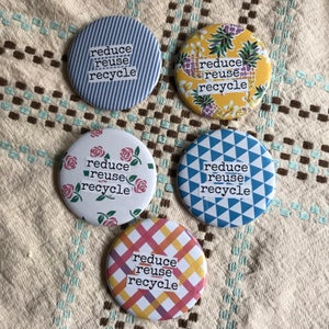 May include: Five round buttons with the words "reduce, reuse, recycle" in a white, blocky font. The buttons have various patterns: blue stripes, yellow pineapples, pink roses, blue triangles, and a colorful plaid.