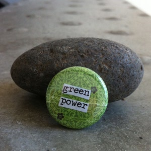 Könnte beinhalten: Ein grüner Knopf mit weißem Hintergrund und dem Aufdruck "green power". Der Knopf liegt auf einem grauen Stein.