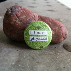May include: A green button with a white background and black text that reads "i heart physics". The button has a floral pattern and is sitting on a brown rock.