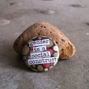 May include: A round button with a brown and beige background with red and yellow polka dots. The button has white text that reads "gender is a social construct".