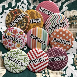 May include: Nine round buttons with different colorful patterns and the word "RESIST." printed in black on each button.