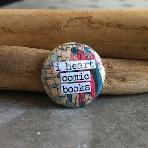 Puede incluir: Un botón redondo con un fondo de cómic que presenta a un superhéroe. El botón tiene el texto "i heart comic books" impreso en él.