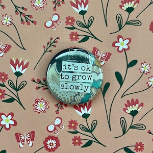 May include: A round button with a floral pattern and the text "it's ok to grow slowly" on a pink background with a floral pattern.
