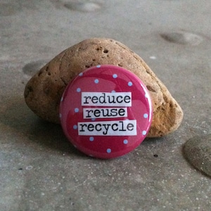 May include: A round, red button with light blue polka dots and the words "reduce reuse recycle" in white block letters. The button is resting on a gray surface next to a brown, textured rock.