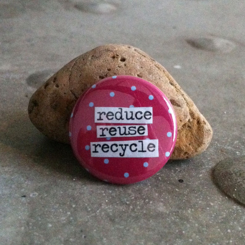 May include: A round, red button with light blue polka dots and the words "reduce reuse recycle" in white block letters. The button is resting on a gray surface next to a brown, textured rock.
