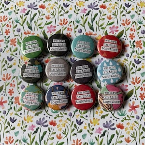 May include: Twelve round buttons with a floral background. Each button has a different patterned design and the text "We are in this together" printed on it.