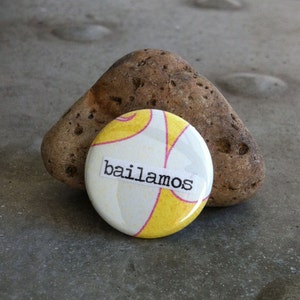 May include: A white button with yellow and pink swirls and the word "bailamos" printed in black.