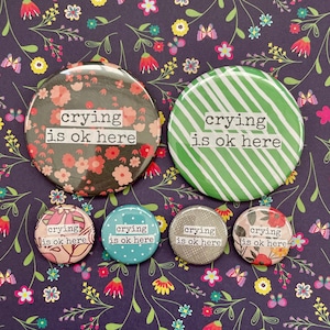 May include: Six round buttons with a white background and black text that reads "crying is ok here". The buttons have different designs, including floral, striped, and speckled patterns.
