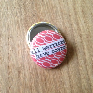 Peut inclure: Un petit miroir compact rond de couleur rouge et blanche à motifs avec une citation qui dit "tous les guerriers ont des cicatrices".