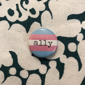 Può includere: Un bottone blu, rosa e bianco con la parola "ally" stampata in nero.
