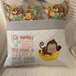 May include: A white quilted pillow with a handle, featuring a colorful jungle animal print and the text "Little monkey, it's time for bed, let's read a story and rest your head." An embroidered monkey rests on a banana.