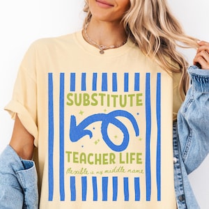 May include: A pale yellow short-sleeve t-shirt featuring a blue vertical striped background. The graphic includes the text "SUBSTITUTE TEACHER LIFE" in green, a blue curved arrow, and the script text "flexible is my middle name" in green.