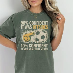 May include: Olive green t-shirt with a soccer-themed graphic. The design features a soccer ball, a referee whistle, and text that reads "90% CONFIDENT IT WAS OFFSIDES 10% CONFIDENT I KNOW WHAT THAT MEANS."