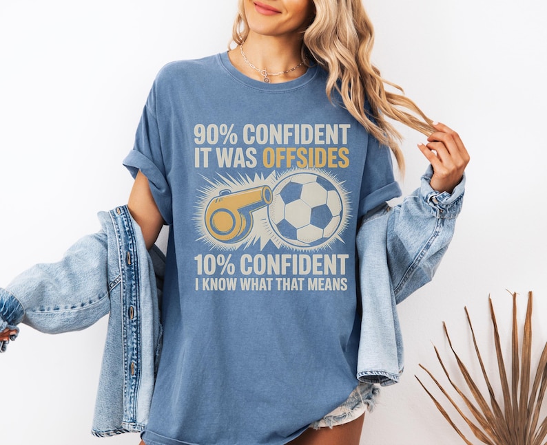 May include: Blue t-shirt with the text "90% CONFIDENT IT WAS OFFSIDES" and "10% CONFIDENT I KNOW WHAT THAT MEANS". The shirt features a soccer ball and a whistle graphic in yellow and white.