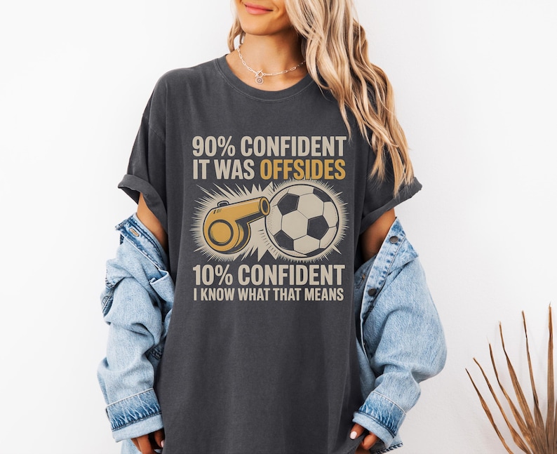 May include: Dark gray t-shirt with a soccer theme. The shirt features a graphic of a soccer ball and a whistle, along with the text "90% CONFIDENT IT WAS OFFSIDES" and "10% CONFIDENT I KNOW WHAT THAT MEANS."