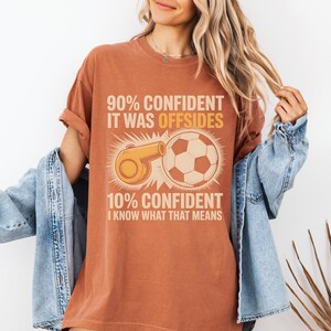 May include: A rust-colored t-shirt with a soccer-themed graphic. The graphic includes a soccer ball, a whistle, and text that reads "90% CONFIDENT IT WAS OFFSIDES" and "10% CONFIDENT I KNOW WHAT THAT MEANS."