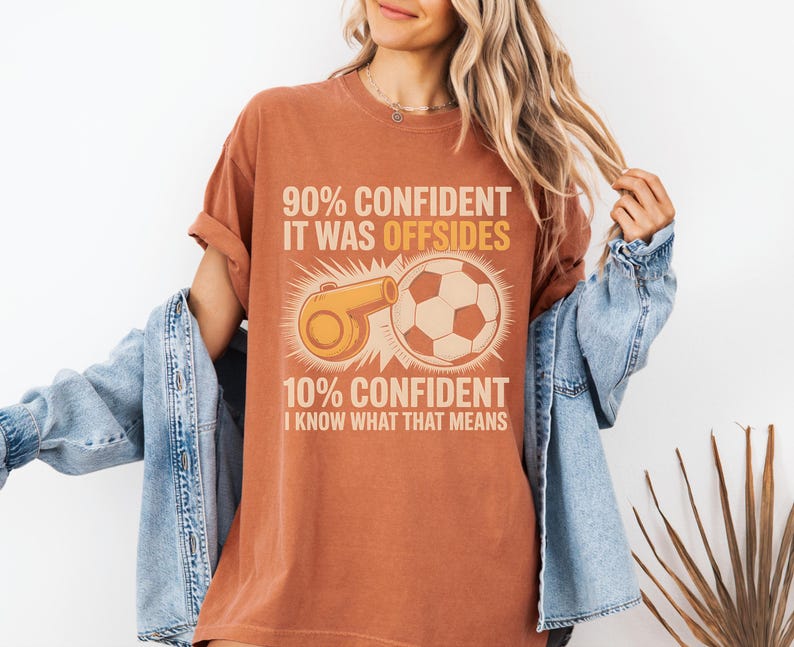 May include: A rust-colored t-shirt with a soccer-themed graphic. The graphic includes a soccer ball, a whistle, and text that reads "90% CONFIDENT IT WAS OFFSIDES" and "10% CONFIDENT I KNOW WHAT THAT MEANS."