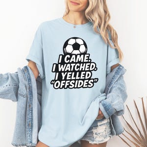 May include: Light blue t-shirt with a soccer ball graphic and the text "I CAME. I WATCHED. I YELLED 'OFFSIDES'". The shirt is made of a soft material.