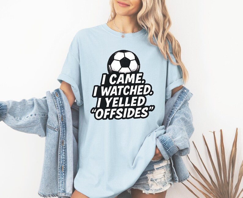 May include: Light blue t-shirt with a soccer ball graphic and the text "I CAME. I WATCHED. I YELLED 'OFFSIDES'". The shirt is made of a soft material.
