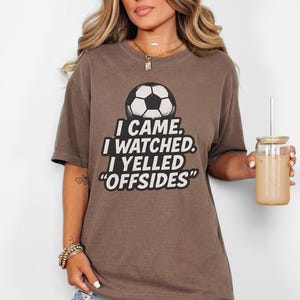 May include: A brown t-shirt with a soccer ball graphic and the text "I CAME. I WATCHED. I YELLED 'OFFSIDES'". The shirt is a casual style, suitable for sports fans.