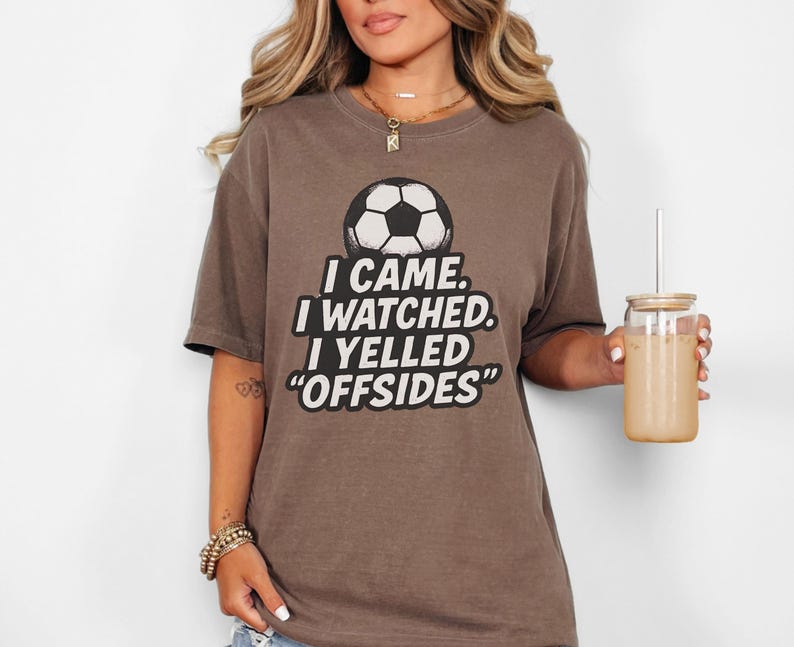 May include: A brown t-shirt with a soccer ball graphic and the text "I CAME. I WATCHED. I YELLED 'OFFSIDES'". The shirt is a casual style, suitable for sports fans.