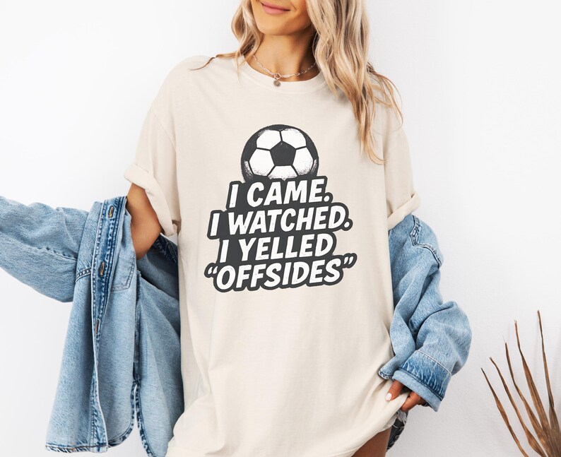 May include: Cream-colored t-shirt with a soccer ball graphic and the text "I CAME. I WATCHED. I YELLED 'OFFSIDES'". The shirt is a relaxed fit and has rolled-up sleeves. A light blue denim jacket is worn over the shirt.